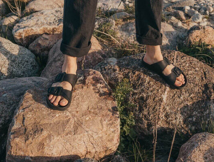 Märt Sandals in Black on rocky ground, showcasing adjustable buckle straps and leather comfort.