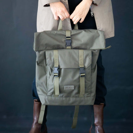 Person holding an olive green NOEL rolltop backpack made in Europe, styled with a taupe jacket and brown boots.