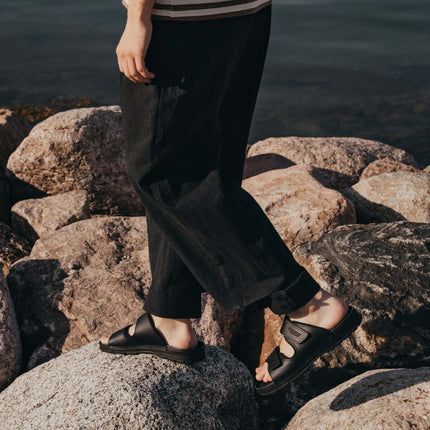 Person in a striped shirt and black pants standing on rocks by the water, wearing stylish black sandals.
