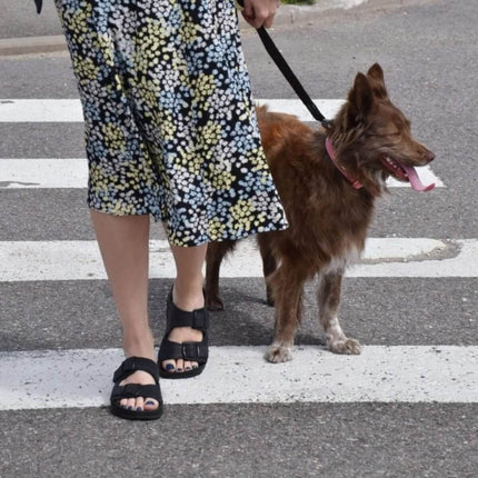 Woman in handmade sandals walking a dog on a crosswalk, showcasing everyday comfort and stylish design.