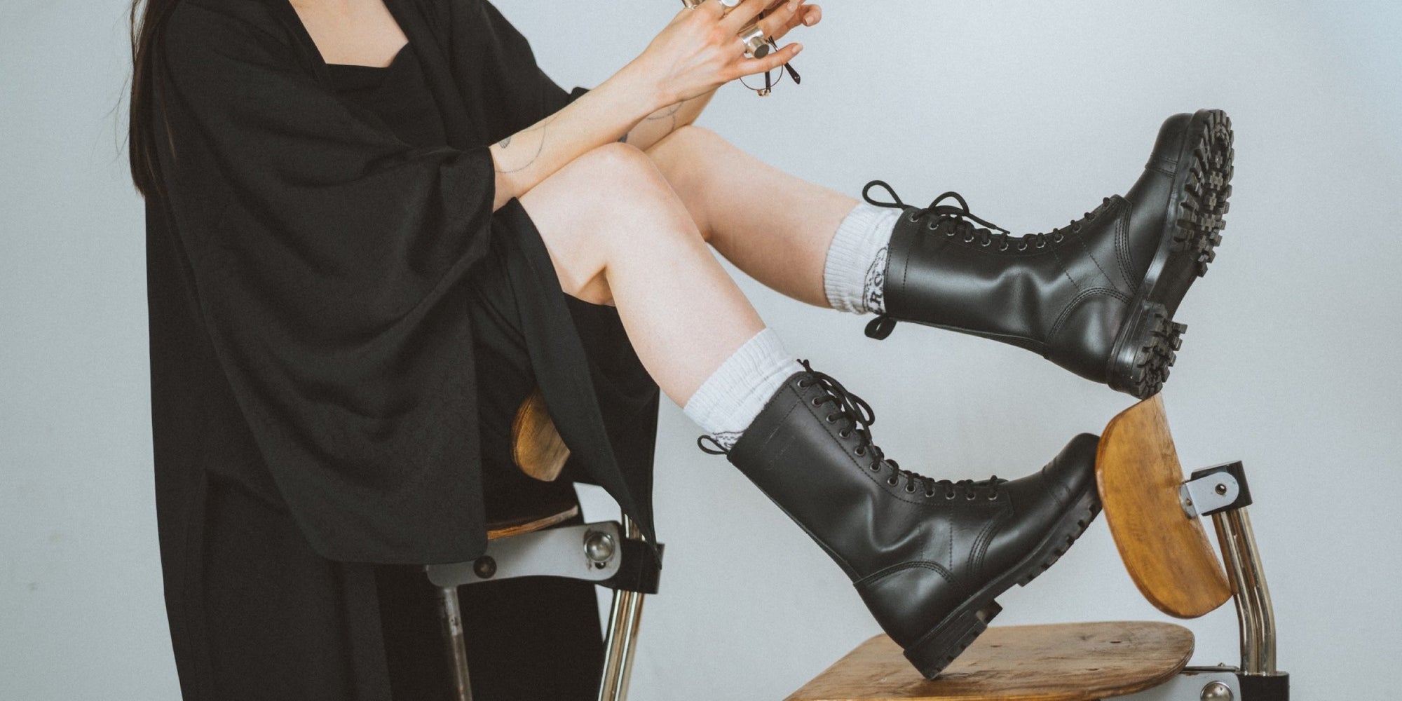 Person wearing black boots sitting on a wooden stool with a plain background