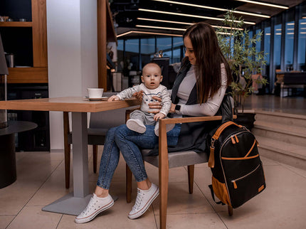Mother sitting with baby at a cafe, featuring a Large Diaper Backpack - Black Coffee beside them.