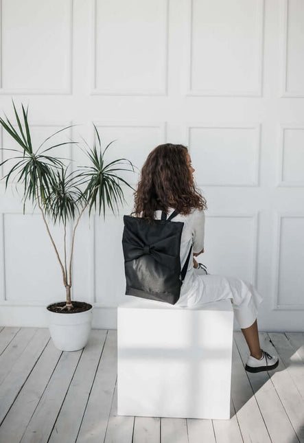 Person sitting with a black waterproof backpack featuring a large bow, surrounded by minimalistic decor.