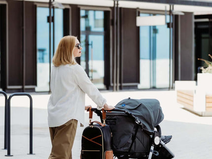 Stylish woman walking with a large black diaper backpack and a stroller in an urban setting.