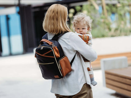 Parent carrying a small black coffee diaper backpack while holding a child in a modern outdoor setting.