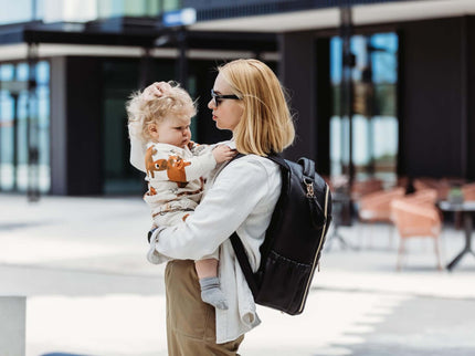 Mom holding child while wearing a large espresso diaper backpack, showcasing style and convenience in an outdoor setting.