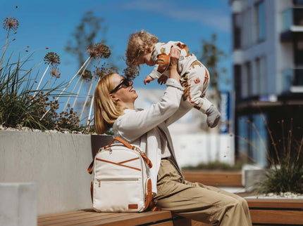 A stylish woman joyfully holds her child while seated next to a small cappuccino diaper backpack outdoors.