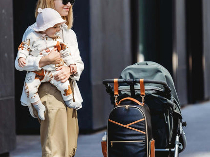 Stylish mother with a baby and large black coffee diaper backpack beside a stroller on a city street.