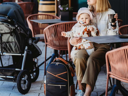 Stylish parent enjoying coffee at cafe with large Black Coffee diaper backpack and child in stroller.