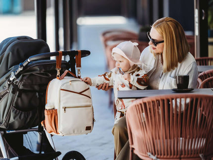 Stylish parent with Small Diaper Backpack in cappuccino color, enjoying coffee with baby in stroller.