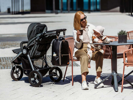 Stylish mother with a baby enjoying coffee at a café, with a large diaper backpack next to a stroller.