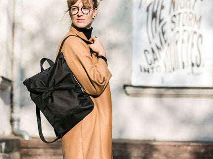 Stylish woman wearing a black waterproof backpack with a bow, standing against a bright urban backdrop.