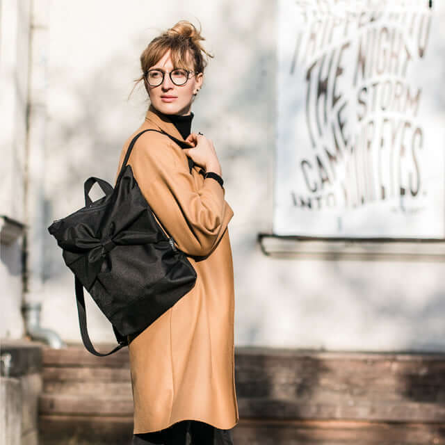 Stylish woman wearing a black waterproof backpack with a bow, standing against a bright urban backdrop.