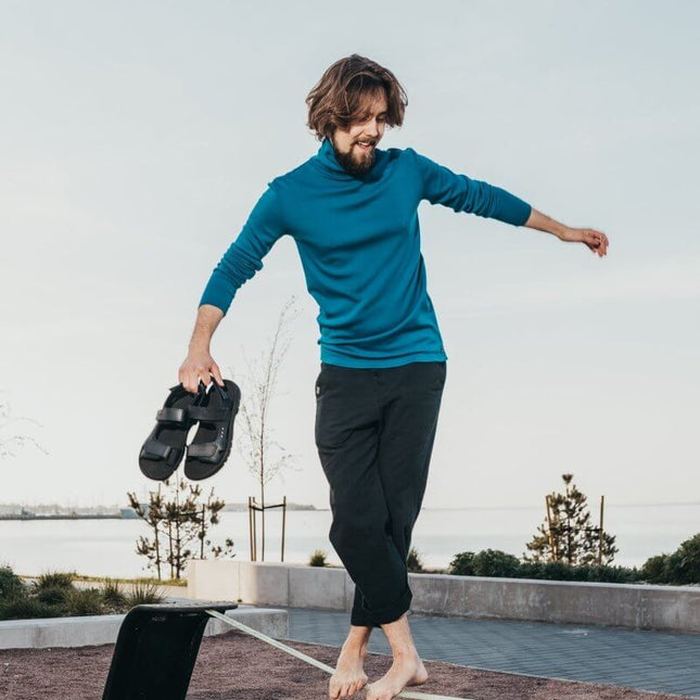 Man balancing on a slackline outdoors, holding Lauri leather sandals, wearing a blue long-sleeve top.