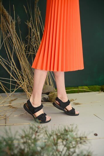 Woman wearing Liiva Women's Leather Sandals in black with a pleated orange skirt, showcasing style and comfort.