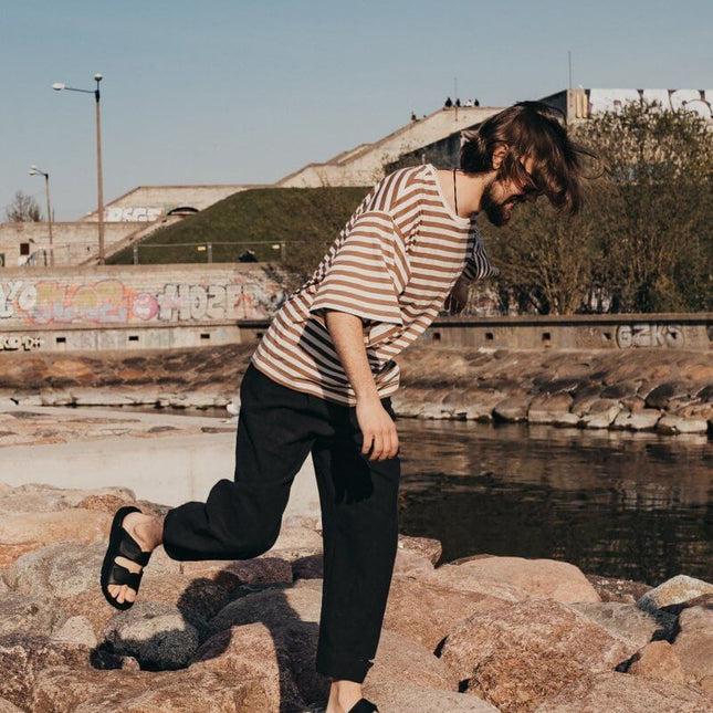 Man wearing Madise Sandals for Him - Black, walking on rocks by a waterway, showcasing casual summer style.