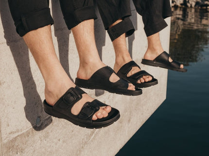 Three pairs of Märt black leather slip-on sandals with adjustable straps, showcasing comfort and style by the water.