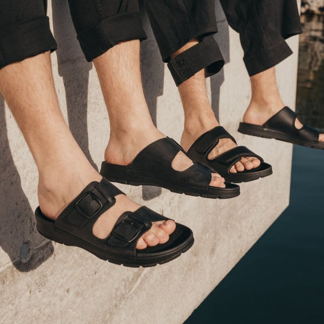 Three pairs of Märt black leather slip-on sandals with adjustable straps, showcasing comfort and style by the water.