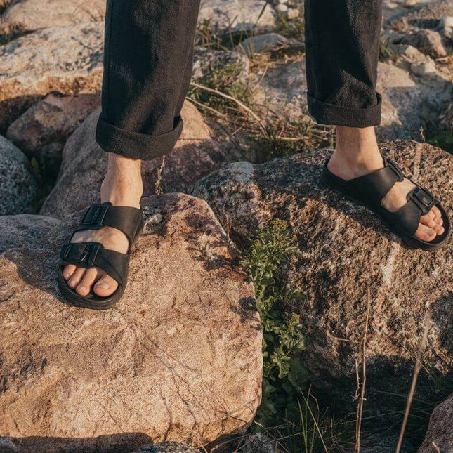 Märt Sandals in Black on rocky ground, showcasing adjustable buckle straps and leather comfort.