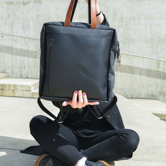 Stockholm Backpack in black, featuring leather handles, held by a person sitting outdoors. Perfect for daily use and travel.