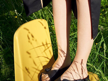 Person wearing TOKU Rome black sandals while standing on a yellow chair in a grassy setting.