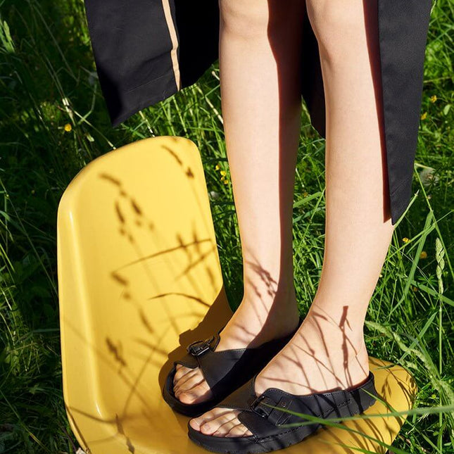 Person wearing TOKU Rome black sandals while standing on a yellow chair in a grassy setting.