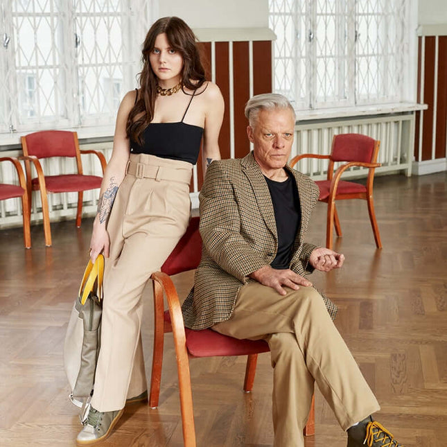 A young woman and an older man posing together in a stylish interior setting, both wearing fashionable outfits.